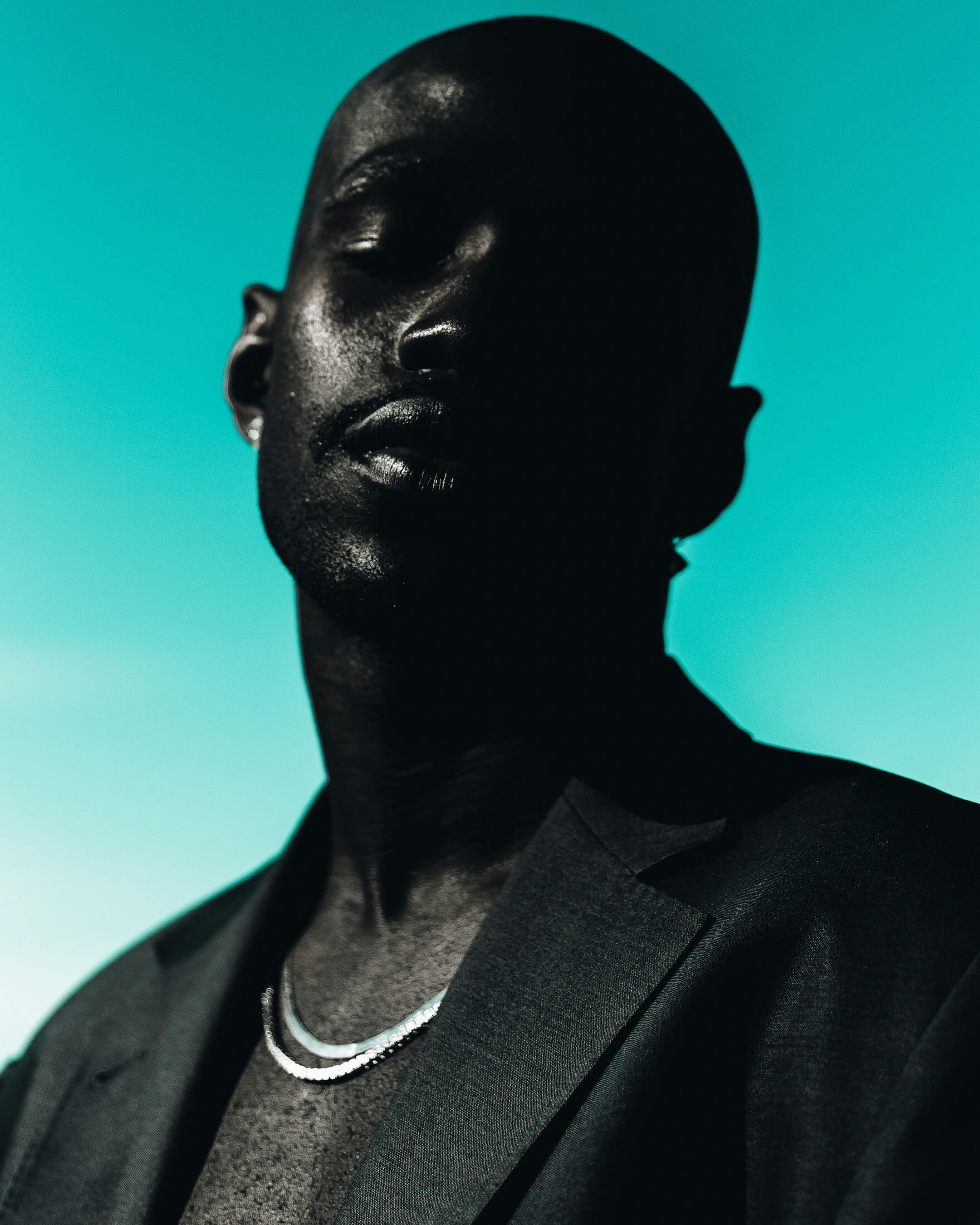Porträtt av en person med rakad frisyr som blickar mot kameran. Personen bär en mörk kavaj och ett halsband, och är fotograferad mot en turkos bakgrund. Det riktade ljuset skapar starka kontraster mellan ljus och skugga och ger bilden ett stilrent och dramatiskt uttryck.