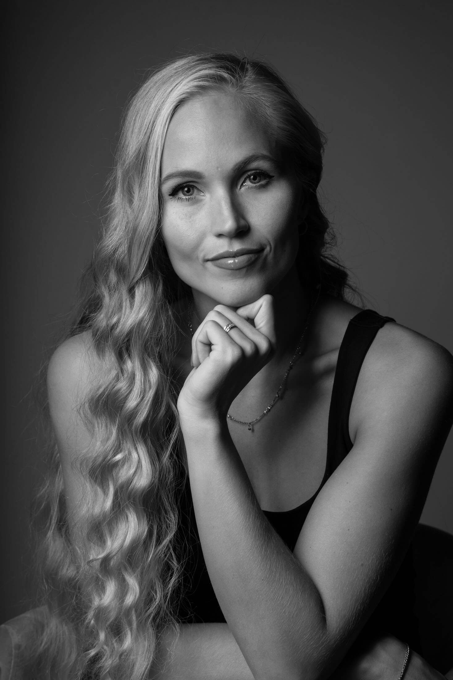 Svartvitt porträtt från midjan upp taget av fotograf i studio där person med långt vågigt hår sitter med hand vilande under hakan. Personen blickar mot kameran med ett lugnt och självsäkert uttryck. Det mjuka studioljuset framhäver ansiktsdrag, hår och händer mot en mörk bakgrund.