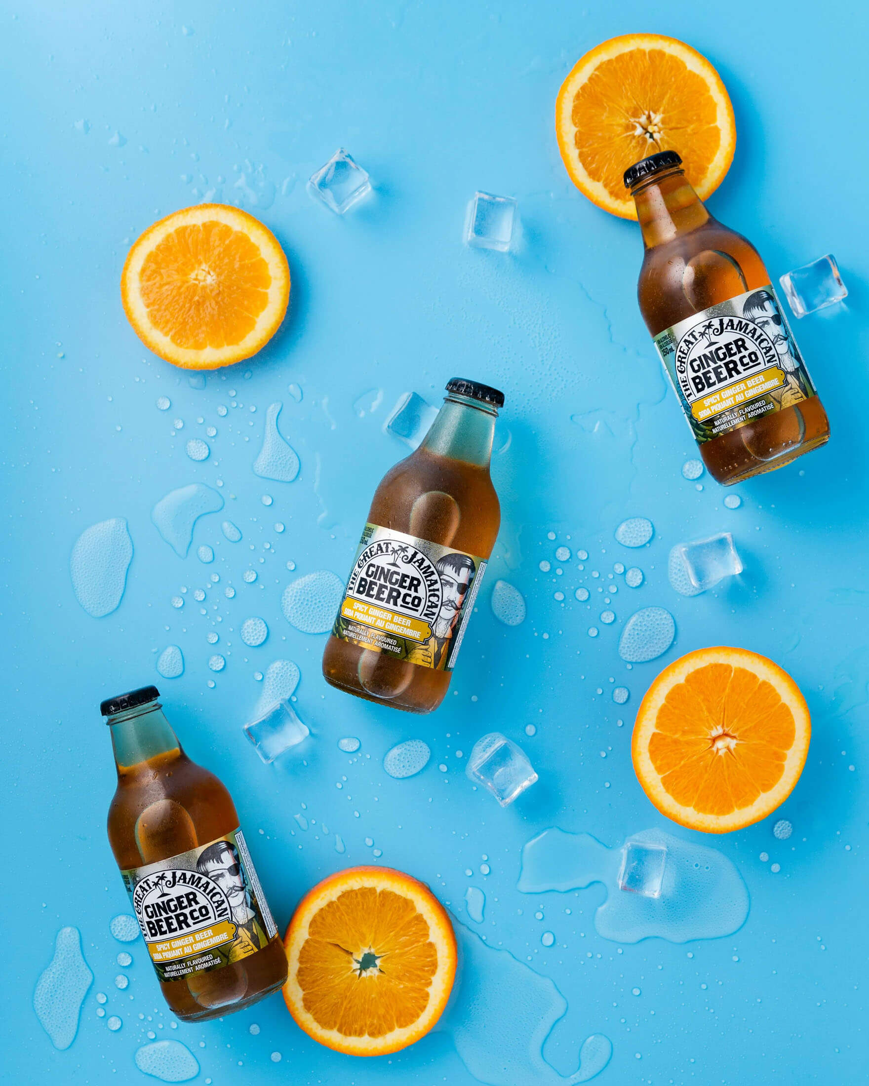 Fotografering i studio i Stockholm där flaskor med ginger beer ligger arrangerade på en blå bakgrund tillsammans med apelsinskivor, isbitar och vattendroppar. Den färgstarka kompositionen framhäver dryckens fräscha karaktär och skapar ett professionellt reklamfoto för marknadsföring och produktkampanjer.