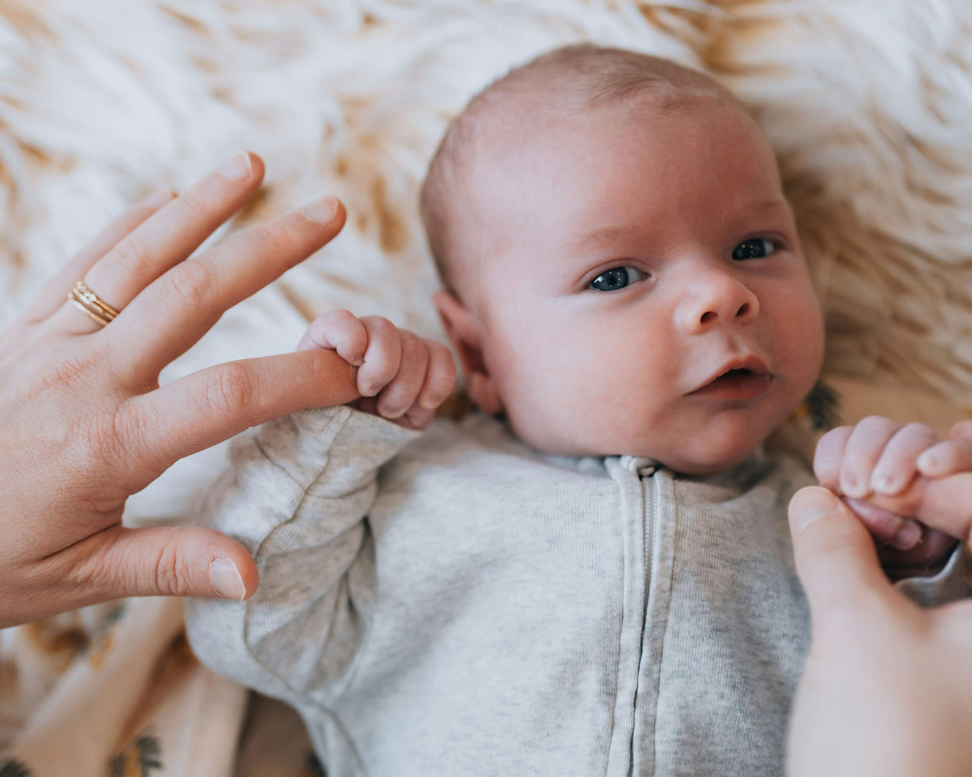 Ett spädbarn ligger på en mjuk, ljus filt och tittar mot kameran med ett lugnt, nyfiket och uppmärksamt uttryck. Barnet bär en ljus heldräkt och håller en vuxen persons finger med båda händerna. Bilden är tagen på nära håll.