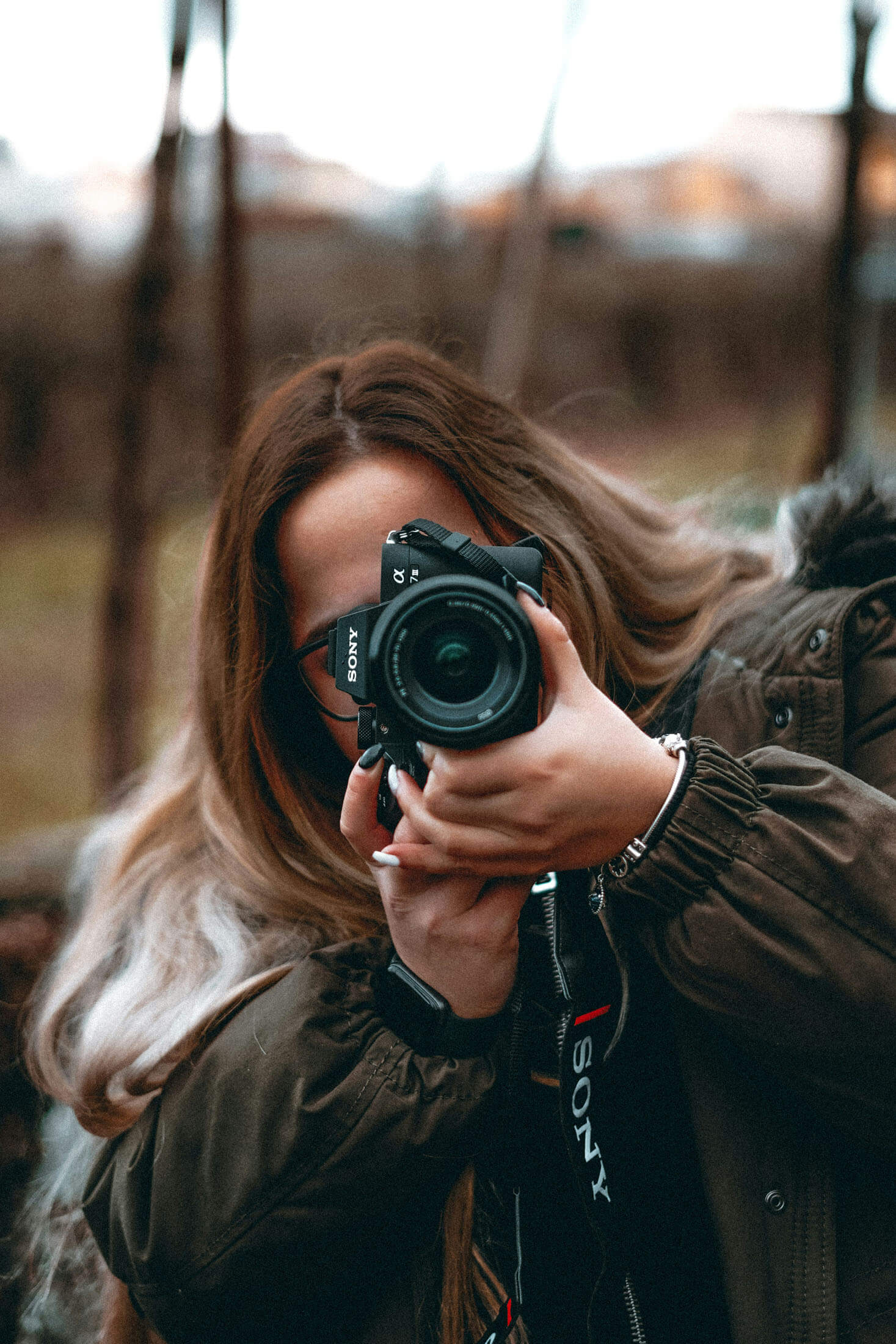 Person håller en systemkamera framför ansiktet och fotograferar utomhus, med kort skärpedjup och suddig bakgrund av natur eller stad.