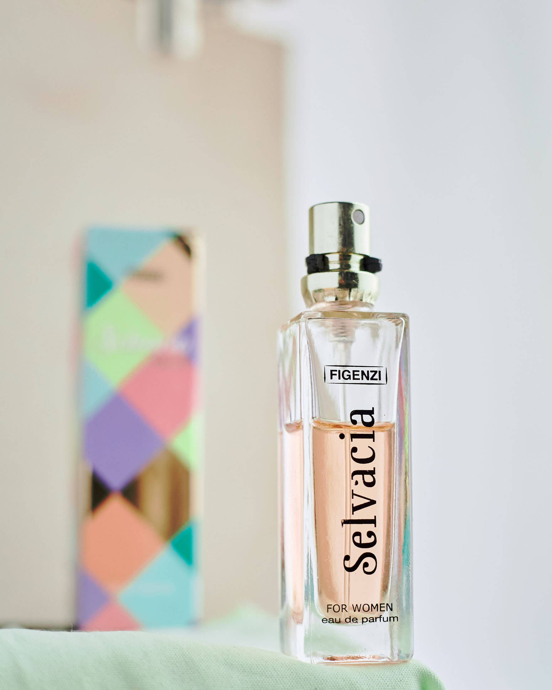Produktfotografering i Stockholm där en parfymflaska i glas presenteras mot ljus bakgrund med en färgglad produktförpackning placerad bakom. Den grunda skärpan och den professionella ljussättningen framhäver produktens design, material och premiumkänsla i en stilren produktbild för webbshop och reklam.