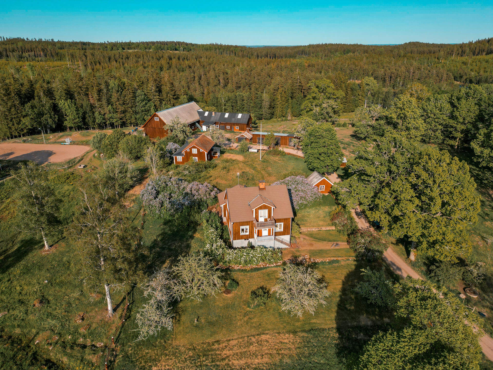 Flygbild över en lantlig fastighet med flera röda byggnader omgivna av träd och grönska. En grusväg leder genom området, och skog breder ut sig i bakgrunden. Bilden ger en överblick över den naturnära miljön och den omgivande landsbygden