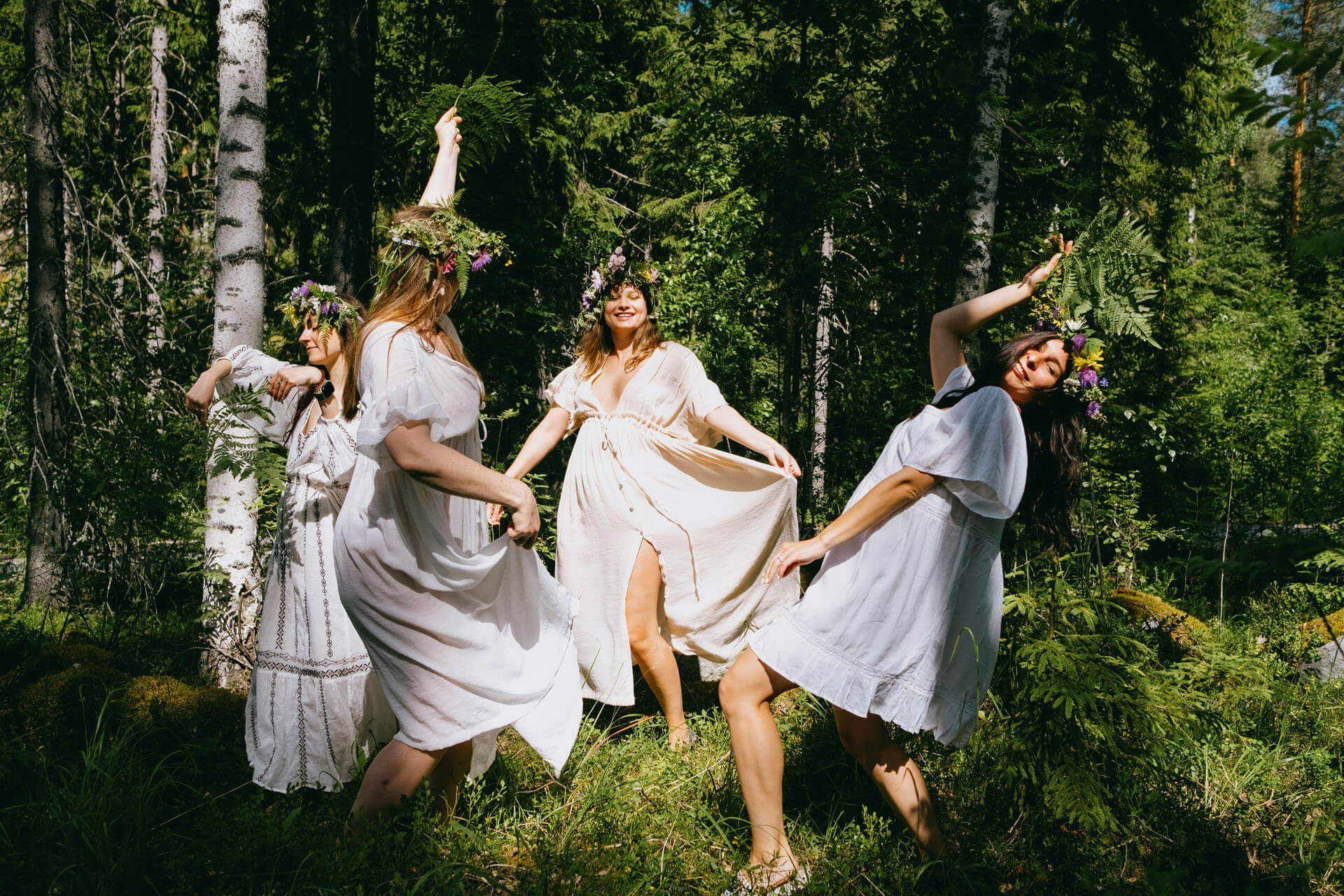 Fyra brudtärnor med blomsterkransar dansar tillsammans i skogen med vita klänningar.