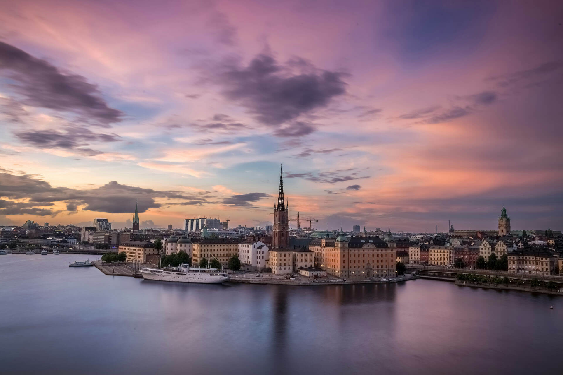Panorama över Stockholm i solnedgång med upplysta byggnader längs vattnet och dramatiska moln i himlen.