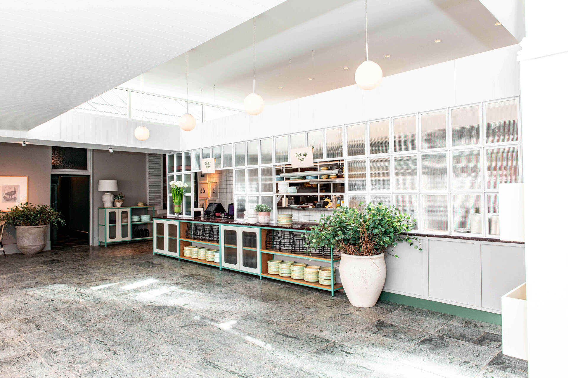 Ljust kök eller caféinteriör med högt tak, stora glaspartier och öppet serveringsområde med hyllor, växter och mjukt dagsljus som flödar in över stengolv.