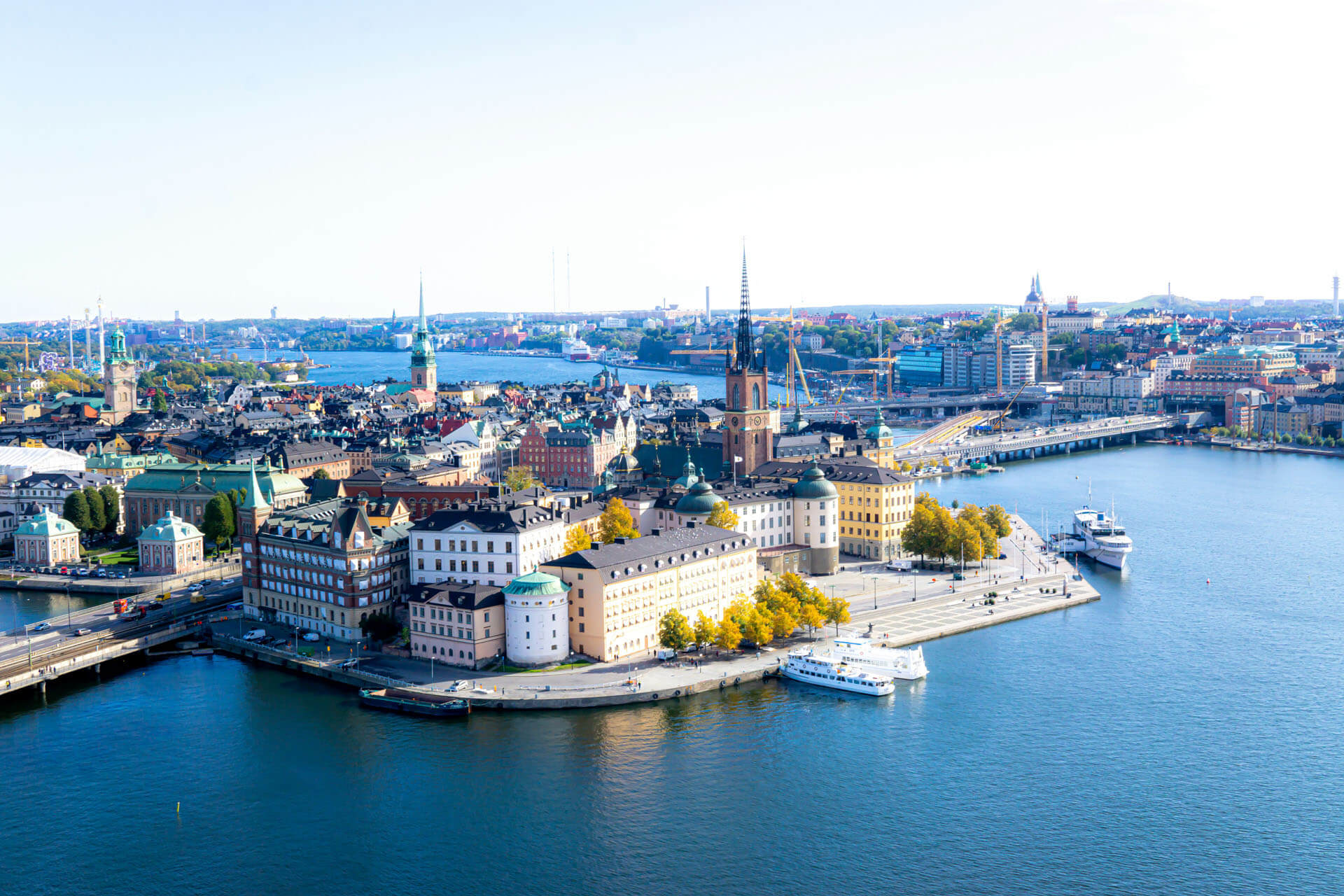 Drönarbild över Gamla stan i Stockholm med kyrktorn, färgglada byggnader och vatten som omger stadskärnan i klart dagsljus.