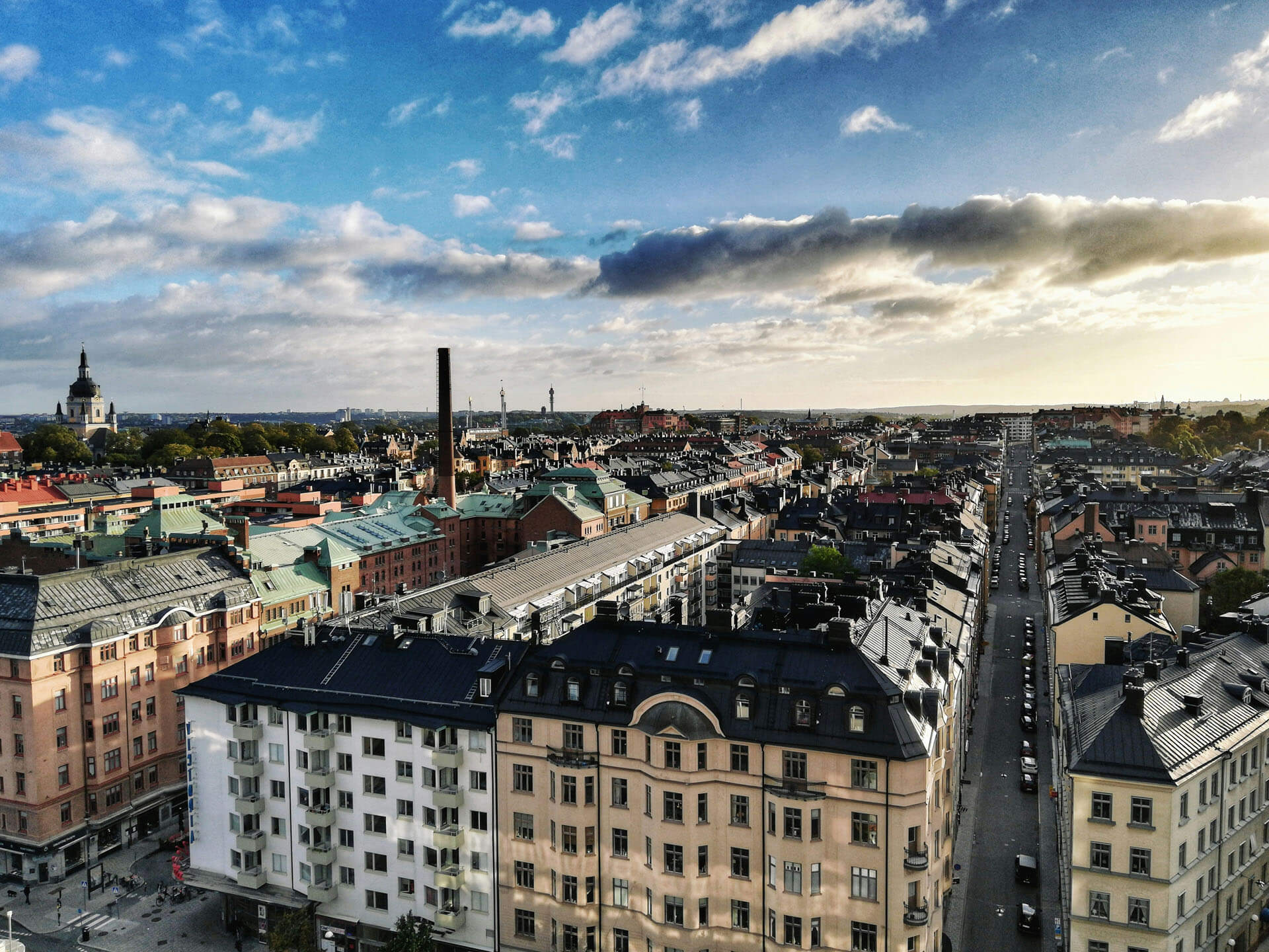 Flygfoto över tät stadsmiljö med äldre bostadshus, lång rak gata och kyrka i bakgrunden, fotograferad i mjukt eftermiddagsljus.