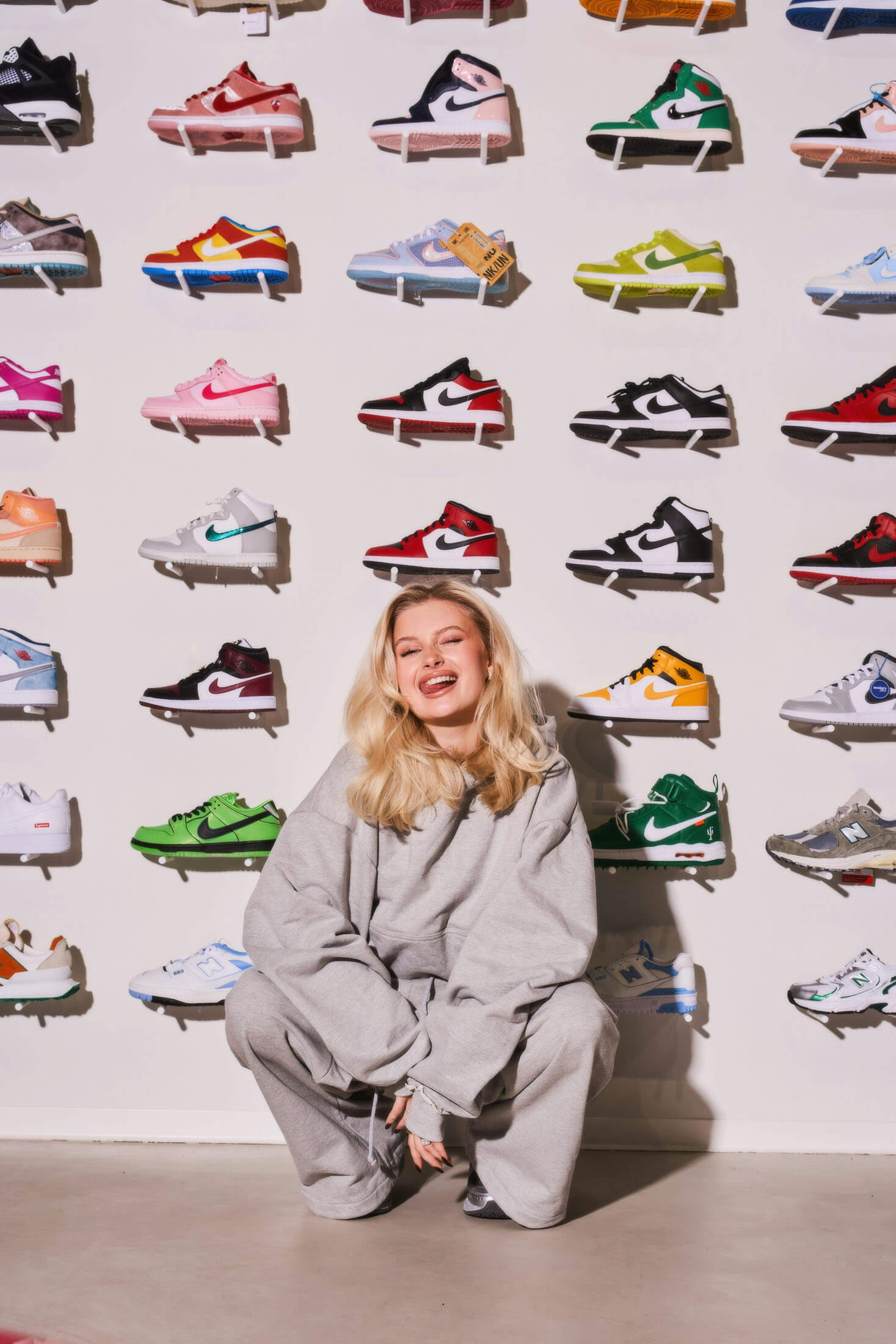 Reklamfotografering i Stockholm där en blond kvinna sitter hukande på golvet framför en vägg fylld med färgglada sneakers uppställda i rader. Med ett leende, en lekfull närvaro skapar hon ett modernt kampanjfoto. Den grafiska bakgrunden och de starka färgerna förstärker uttrycket i en reklambild för streetwear, mode och varumärkeskommunikation.