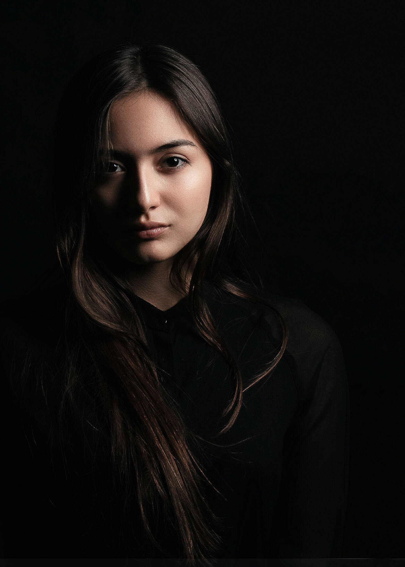 Porträtt av en person med långt mörkt hår som blickar mot kameran med ett lugnt och allvarligt uttryck. Personen bär en mörk tröja och är fotograferad mot en mörk bakgrund. Det mjuka ljuset framhäver ansiktet och skapar en stämningsfull bild med stark kontrast mellan ljus och skugga.