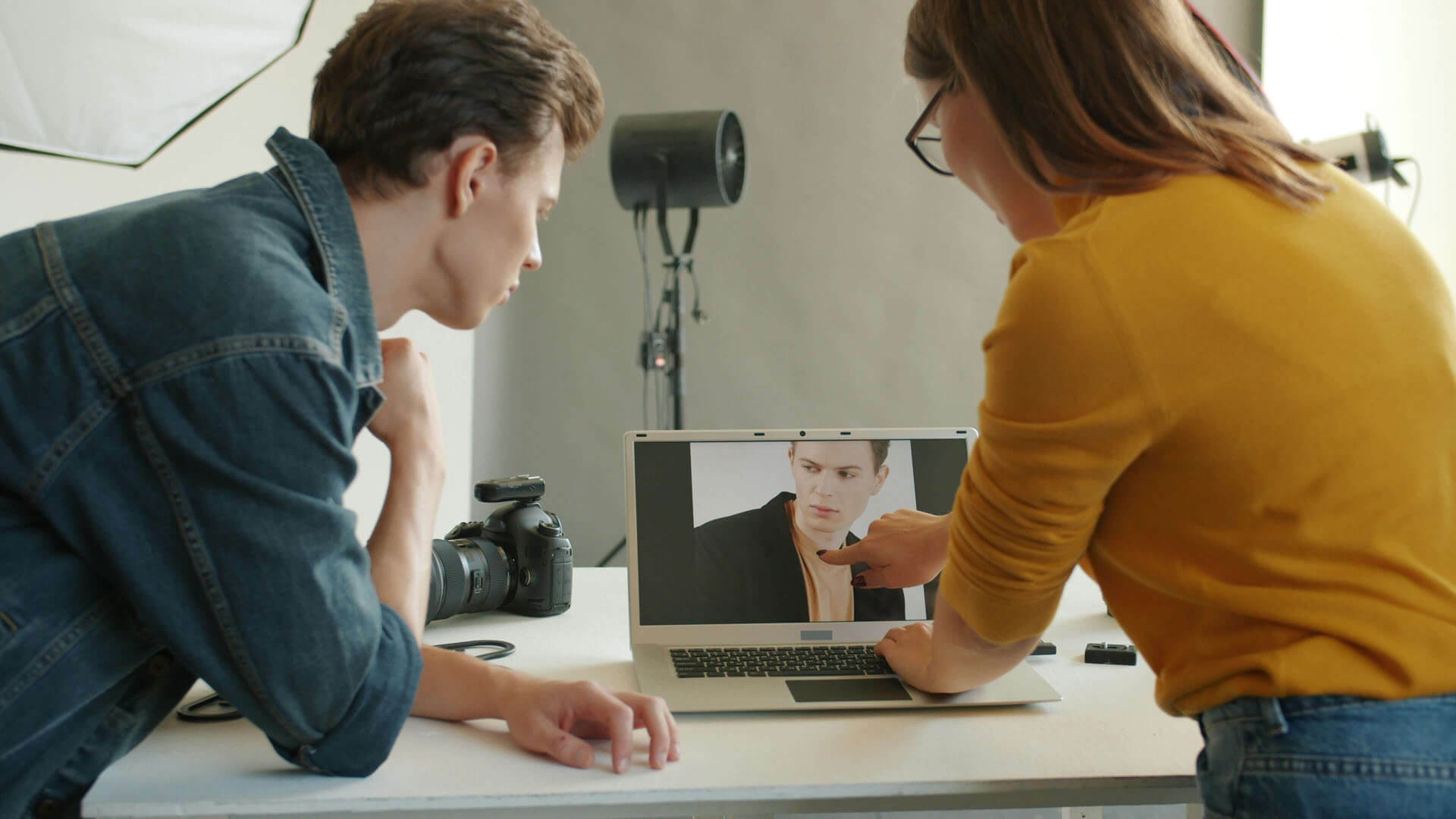 Två personer står vid ett bord i studio och granskar en bild på en laptop, med kamera och studiobelysning synliga i bakgrunden.