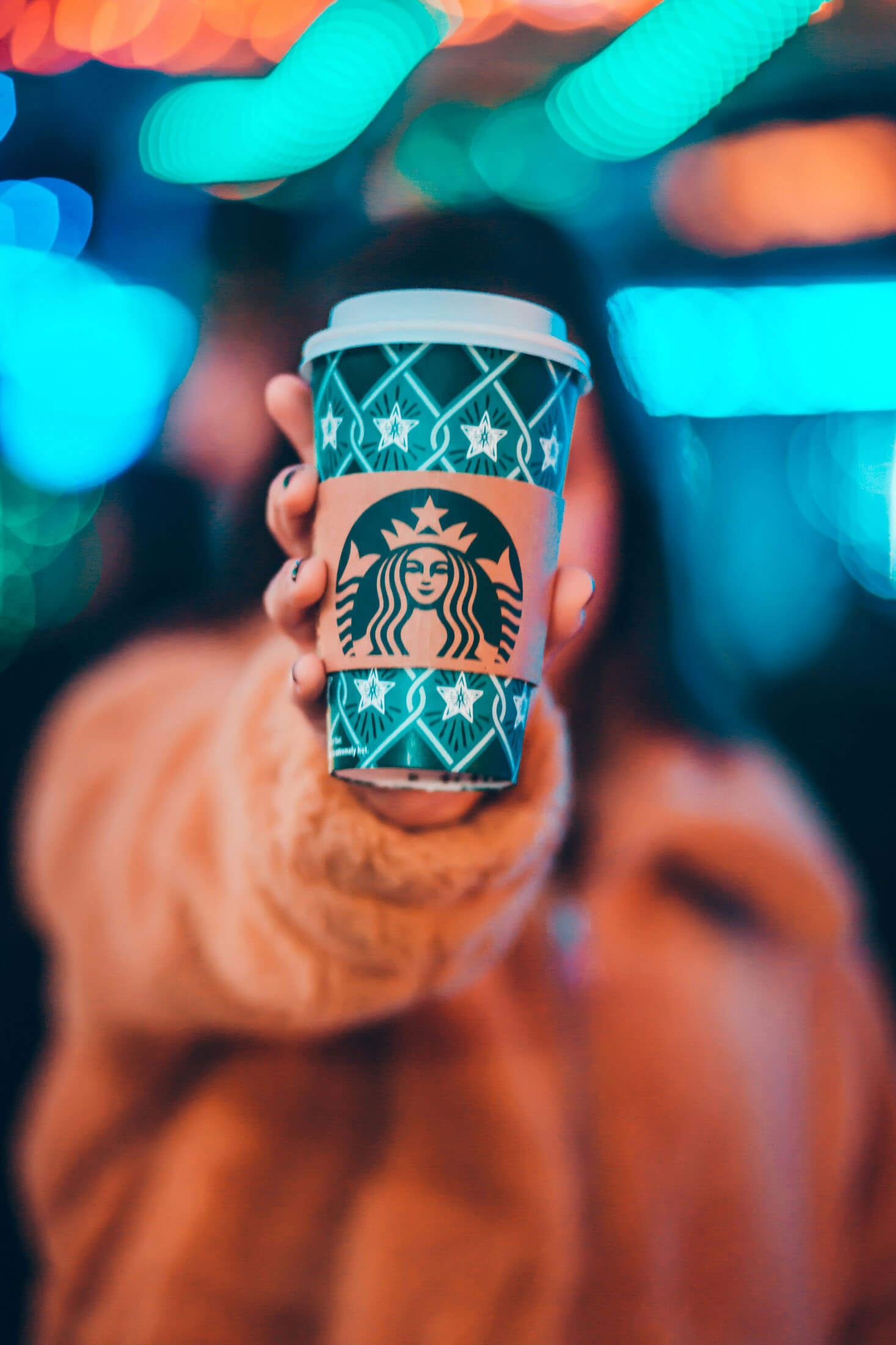 Reklamfoto taget av fotograf i Stockholm där en takeaway-kaffekopp hålls upp i förgrunden medan personen bakom är oskarp. De färgglada ljusen och den grunda skärpan skapar en stämningsfull livsstilsbild för varumärkesfotografering och marknadsföring.